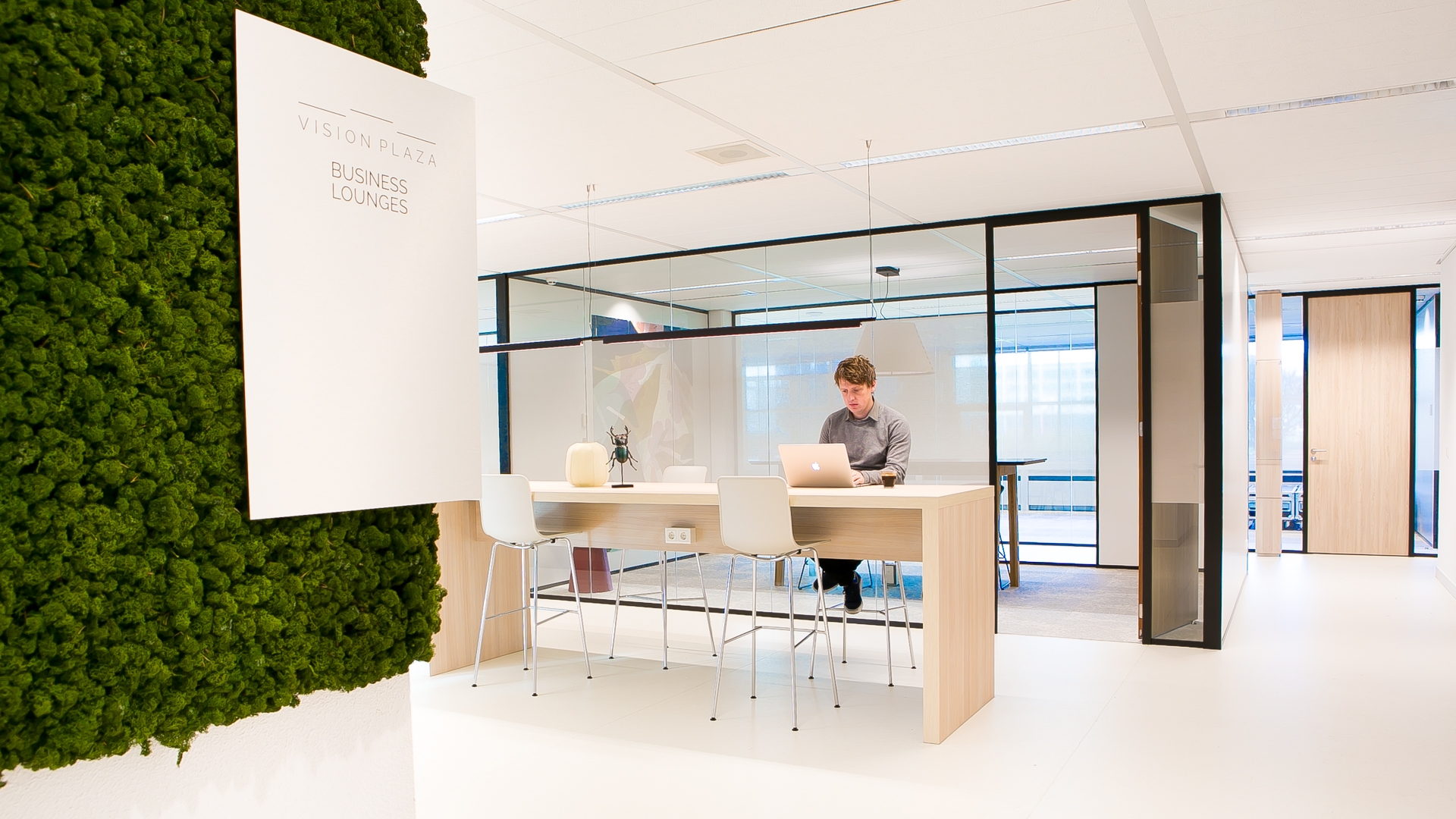 Een moderne kantoorruimte met een hoge tafel en barkrukken waar een persoon achter een laptop werkt. Links is een groene plantenwand zichtbaar met een bord aan de muur. Op de achtergrond bevindt zich een glazen vergaderruimte met zwarte frames.