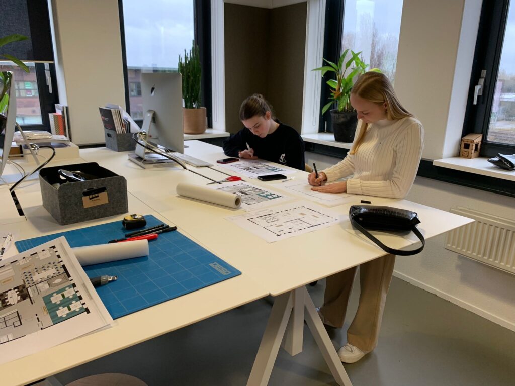 Twee vrouwen zitten aan een bureau in een kantoor en werken geconcentreerd aan bouwtekeningen. Op tafel liggen een snijmat, opgerolde tekeningen, markeerstiften en een opbergbox met kantoorbenodigdheden. Eén vrouw schrijft aantekeningen terwijl de ander d