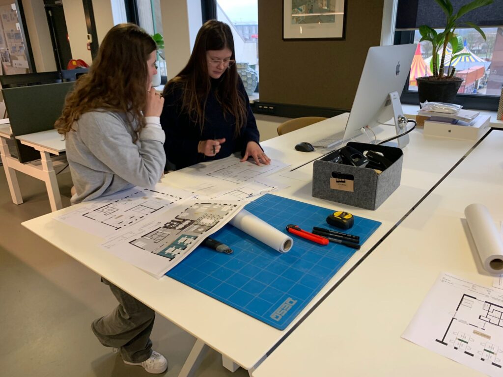Twee vrouwen zitten samen aan een bureau in een kantoor en bekijken bouwtekeningen die op tafel liggen. Op het bureau liggen een snijmat, opgerolde tekeningen, stiften en een opbergbox met kantoorbenodigdheden. Eén van hen wijst naar de tekening terwijl z
