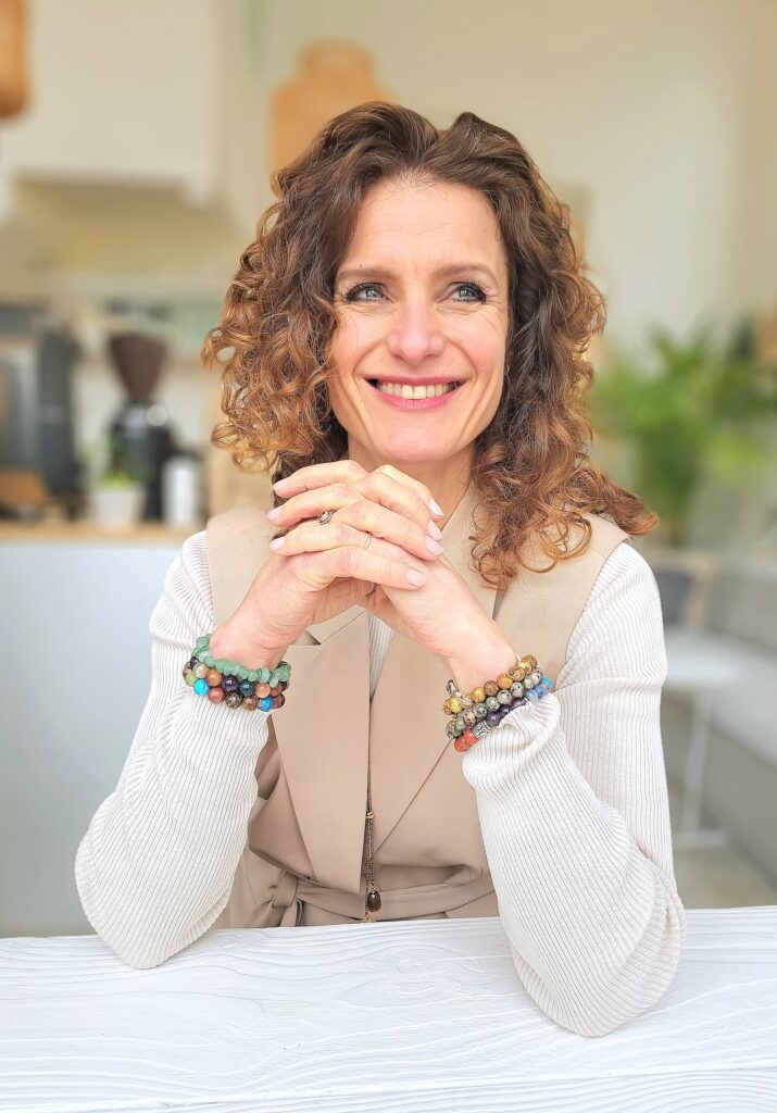 Vrolijk lachende vrouw met krullend haar, zittend aan tafel met haar handen gevouwen, draagster van meerdere kleurrijke armbanden in een lichte, moderne omgeving.