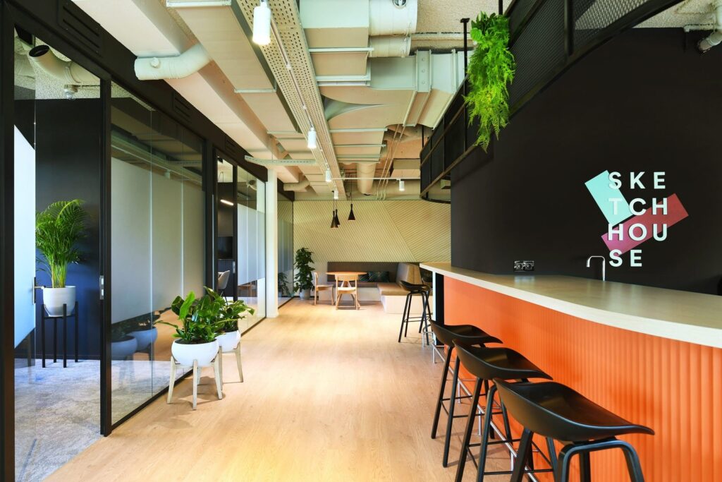 Een moderne kantoorruimte met een lange bar met zwarte barkrukken en een oranje accent aan de onderzijde. Links bevinden zich glazen vergaderruimtes en planten in potten. Het plafond heeft een industriële afwerking met zichtbare leidingen en verlichting.