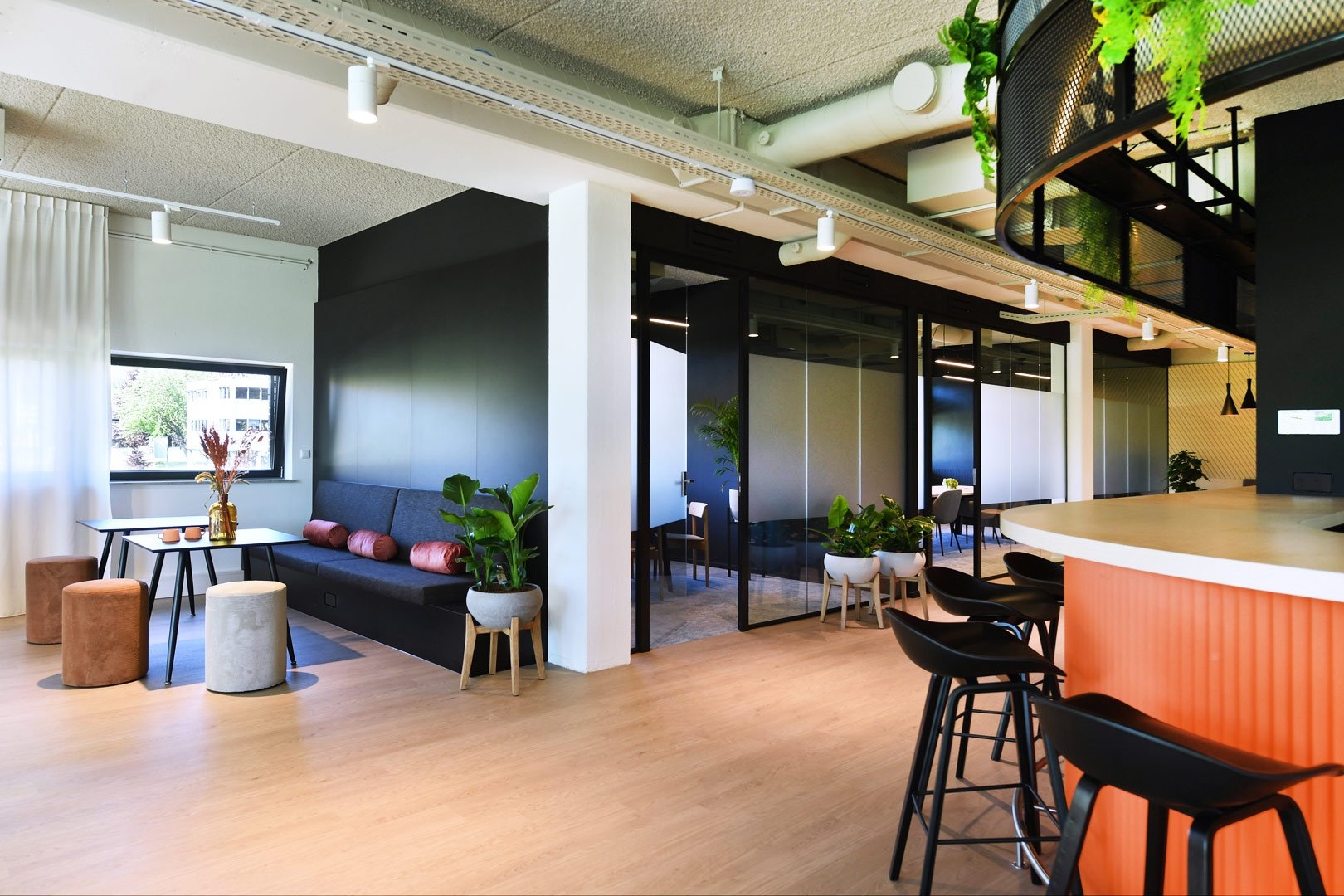 Een moderne kantoorruimte met een bar met zwarte barkrukken en een oranje accent. Links bevindt zich een zithoek met een donkere bank en poefjes bij een raam. In het midden zijn glazen vergaderruimtes met zwarte frames zichtbaar. Het plafond heeft een ind