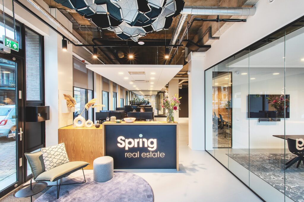 Een moderne kantoorentree met een receptiebalie waarop het logo “Spring real estate” staat. Boven de balie hangt een opvallende designlamp aan een industrieel plafond met zichtbare leidingen. Rechts is een glazen vergaderruimte zichtbaar en de ruimte heef