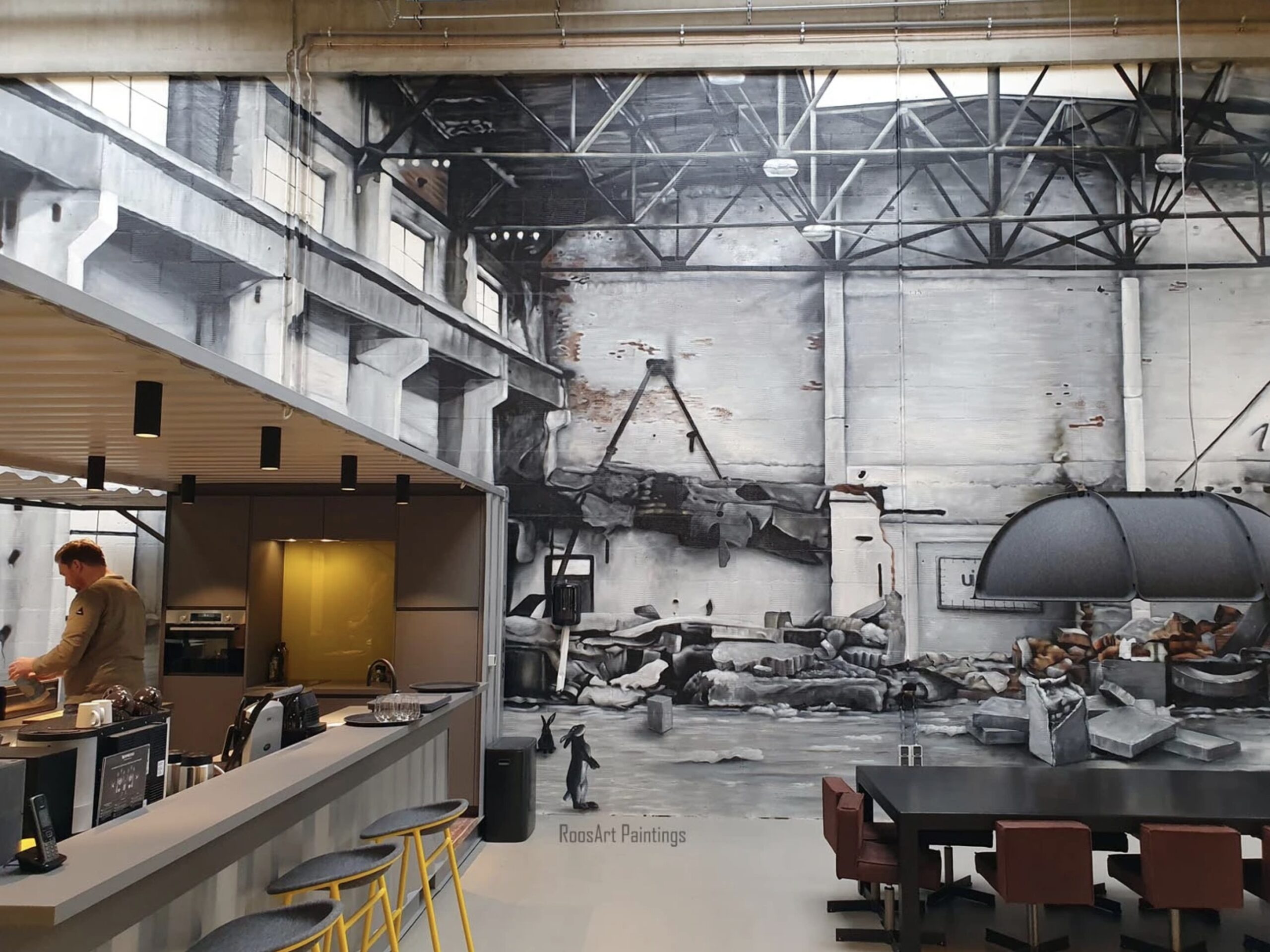 Een ruime, industriële kantoorruimte met een hoge hal en zichtbare stalen constructies aan het plafond. Op de achterwand staat een grote zwart-wit muurschildering van een industriële scène. Links bevindt zich een bar met krukken en een medewerker achter d