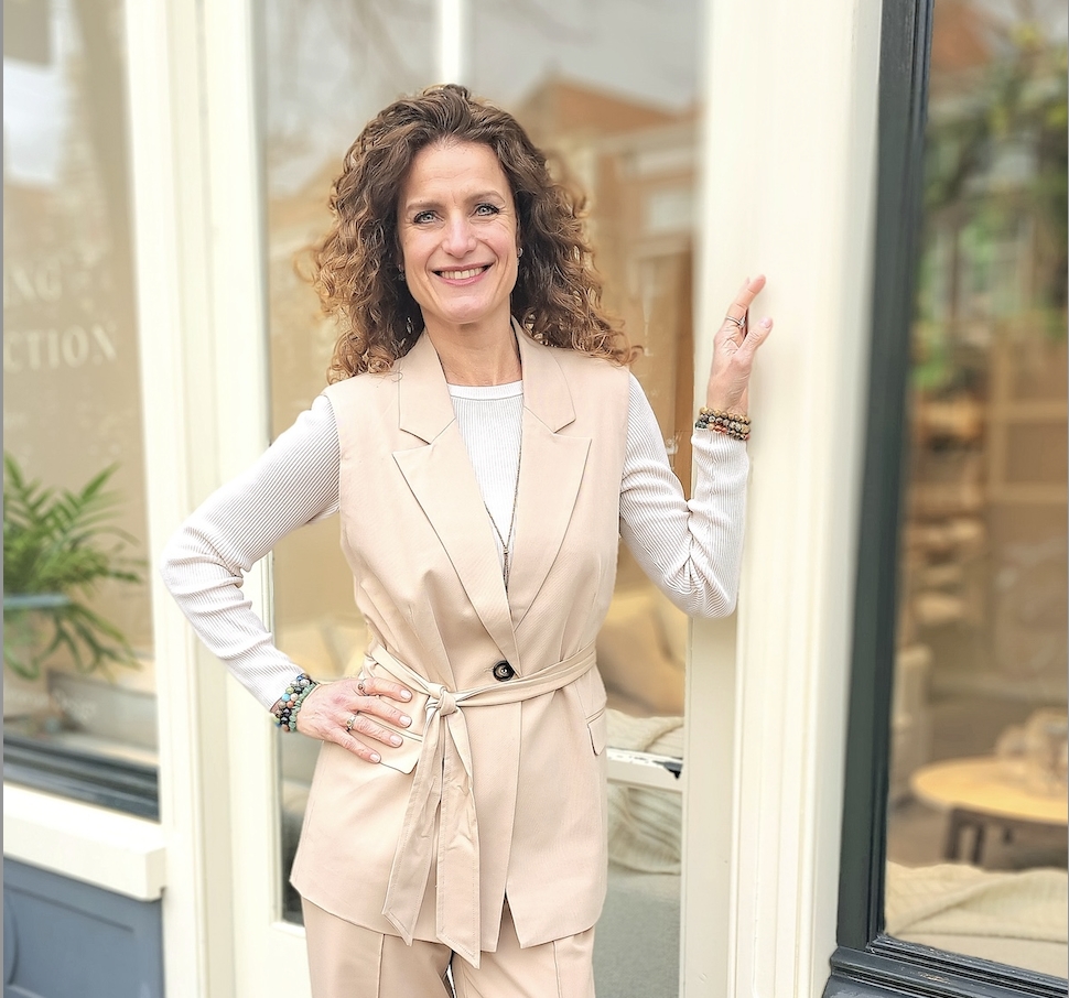 Zelfverzekerd glimlachende vrouw met krullend haar in een beige outfit, staand bij de ingang van een pand met haar hand op de deurpost.