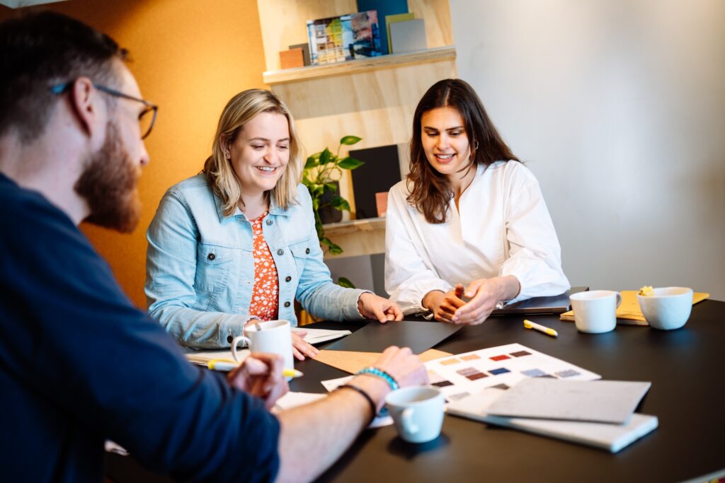 Drie collega’s overleggen aan een tafel met kleurstalen en notities, terwijl ze samenwerken in een informele kantooromgeving met koffiekopjes op tafel.