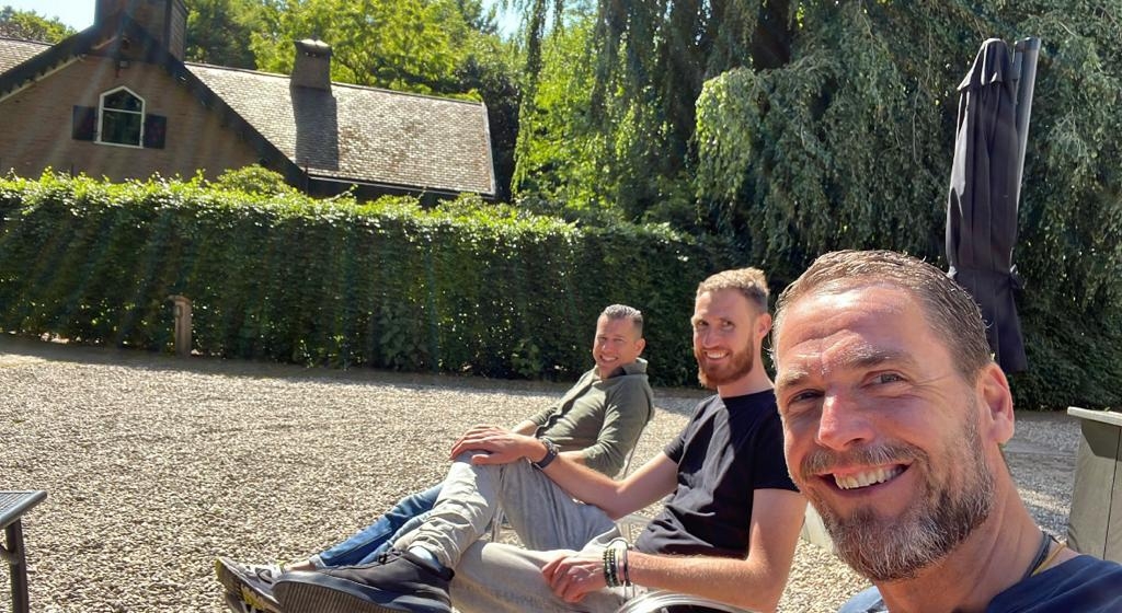 Drie mannen zitten ontspannen naast elkaar op een grindterras in de zon, met op de achtergrond een groene haag, hoge bomen en een landelijk huis met rieten dak. De man op de voorgrond maakt een selfie en glimlacht breed in de camera, terwijl de andere twe