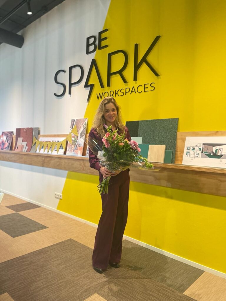 Vrouw met een bos bloemen staat voor een geel muur met het logo van "Be Spark Workspaces".