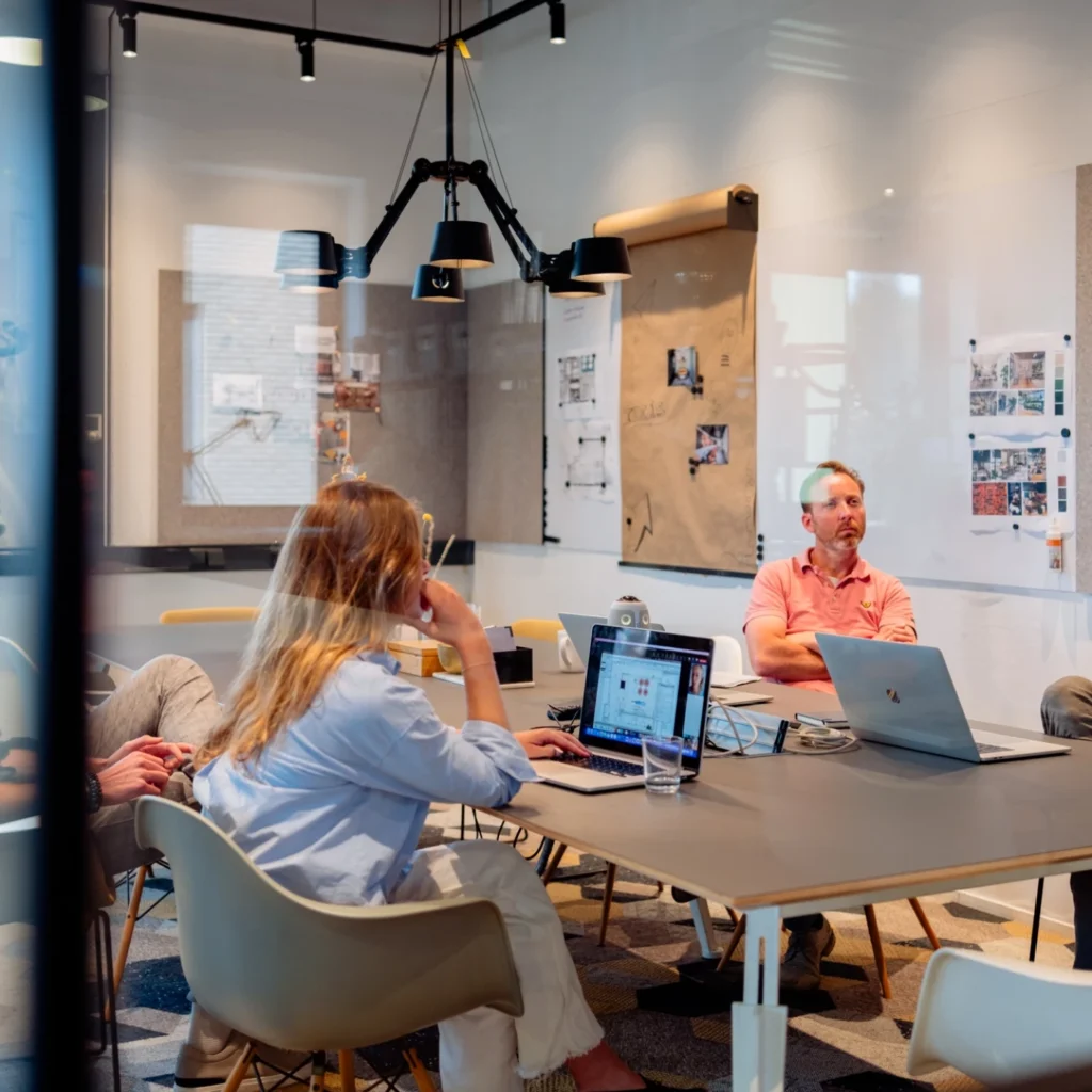 Mensen vergaderen in een moderne kantoorruimte met laptops en presentatiematerialen op de muur.