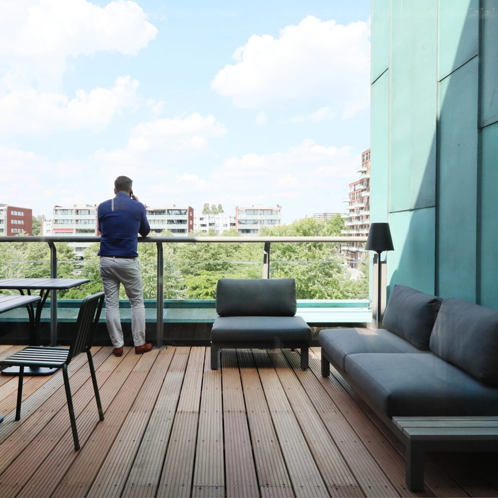 Man staat op een modern kantoor dakterras en kijkt uit over een groene stadsomgeving, met lounge meubels en tafel op houten vlonder.
