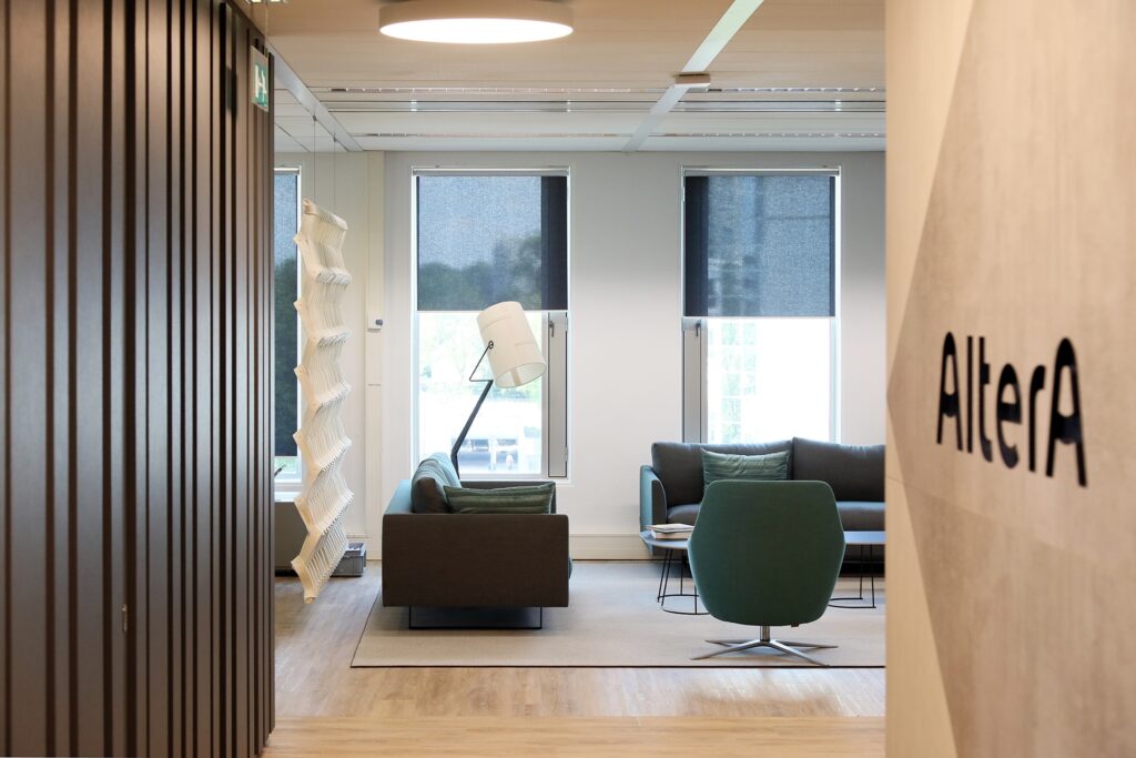 Een moderne zithoek in een kantoor met twee donkere banken en een groene fauteuil rondom een klein rond tafeltje. Grote ramen met half gesloten rolgordijnen zorgen voor daglicht. Links staat een opvallende witte designlamp en de ruimte heeft een lichte ho