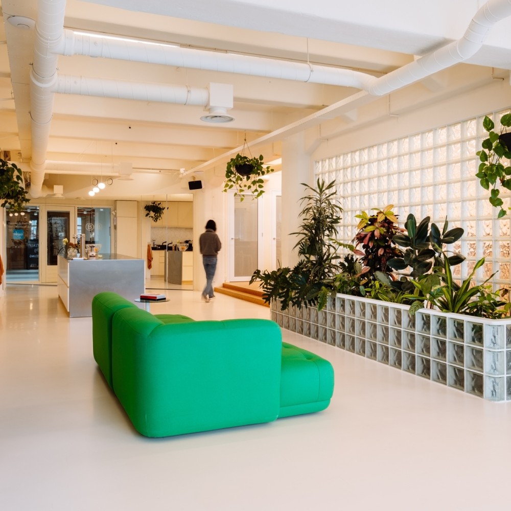 Moderne, lichte kantoorruimte met groene bank, veel planten en een persoon die richting de keuken loopt.