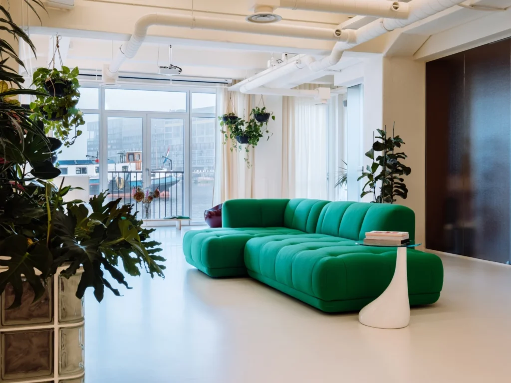 Moderne woonkamer met groene bank en veel hangplanten voor grote ramen met uitzicht op een kanaal.