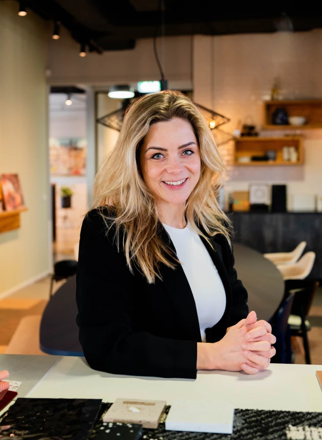 A woman in a black blazer smiles while sitting at a table in a modern office setting.
