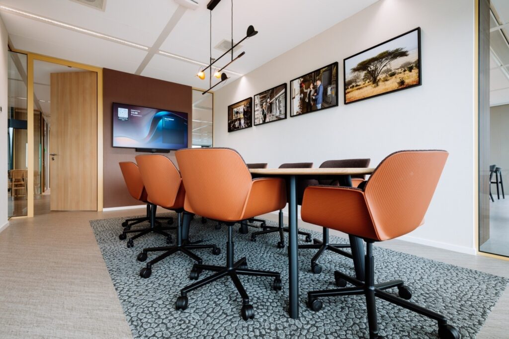 Moderne vergaderruimte met oranje bureaustoelen, houten tafel en groot presentatiescherm aan de muur.
