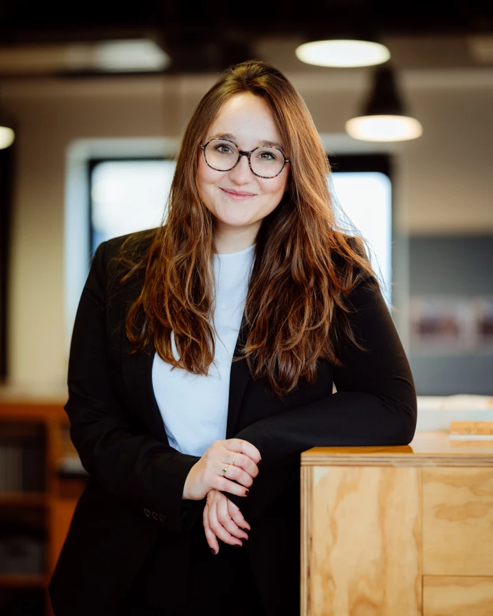 A woman with long hair and glasses is leaning on a wooden surface in an office setting.