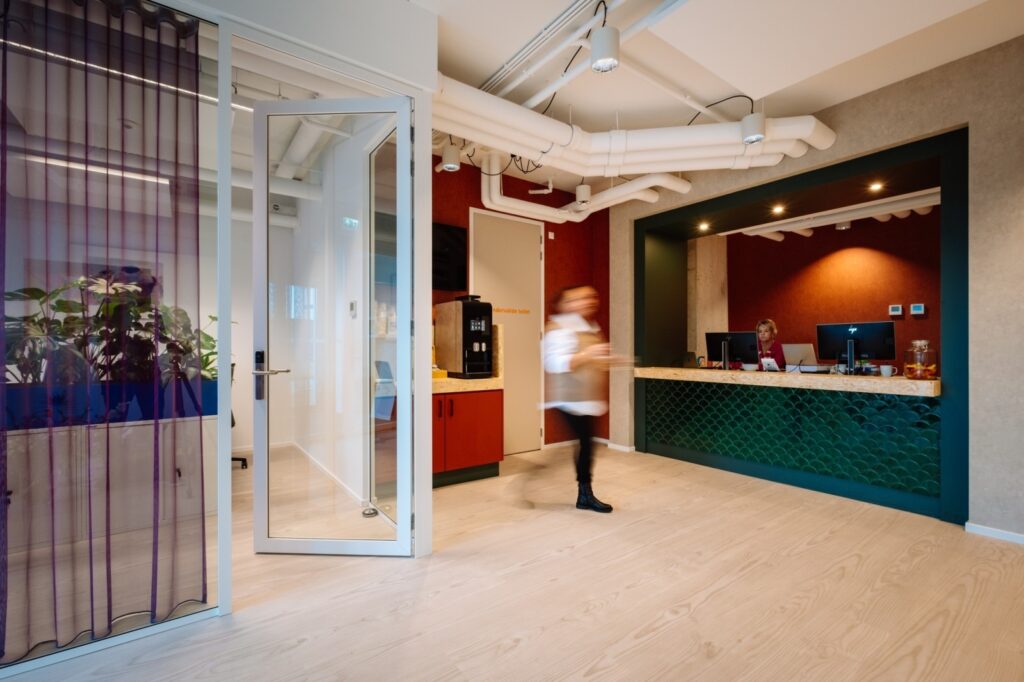 Moderne kantoorentree met open glazen deur, receptiebalie met groene tegels en een medewerker achter de balie, terwijl een persoon door de ruimte loopt.