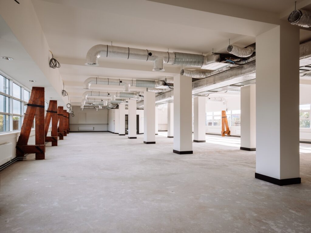 Lege industriële ruimte met ondersteunende houten A-frames en zichtbare ventilatiebuizen aan het plafond.