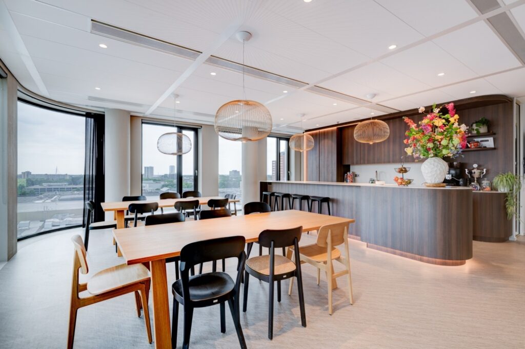 Twee collega’s overleggen aan een hoge tafel in een moderne vergaderruimte met glazen wanden en een betonnen muur. Ze bekijken samen documenten, wat een gefocuste en samenwerkende werksituatie laat zien.