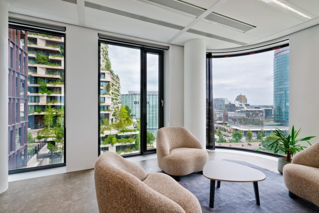 Moderne kantoor lounge met comfortabele stoelen en groot raam met uitzicht op groene architectuur en stadsgebouwen.