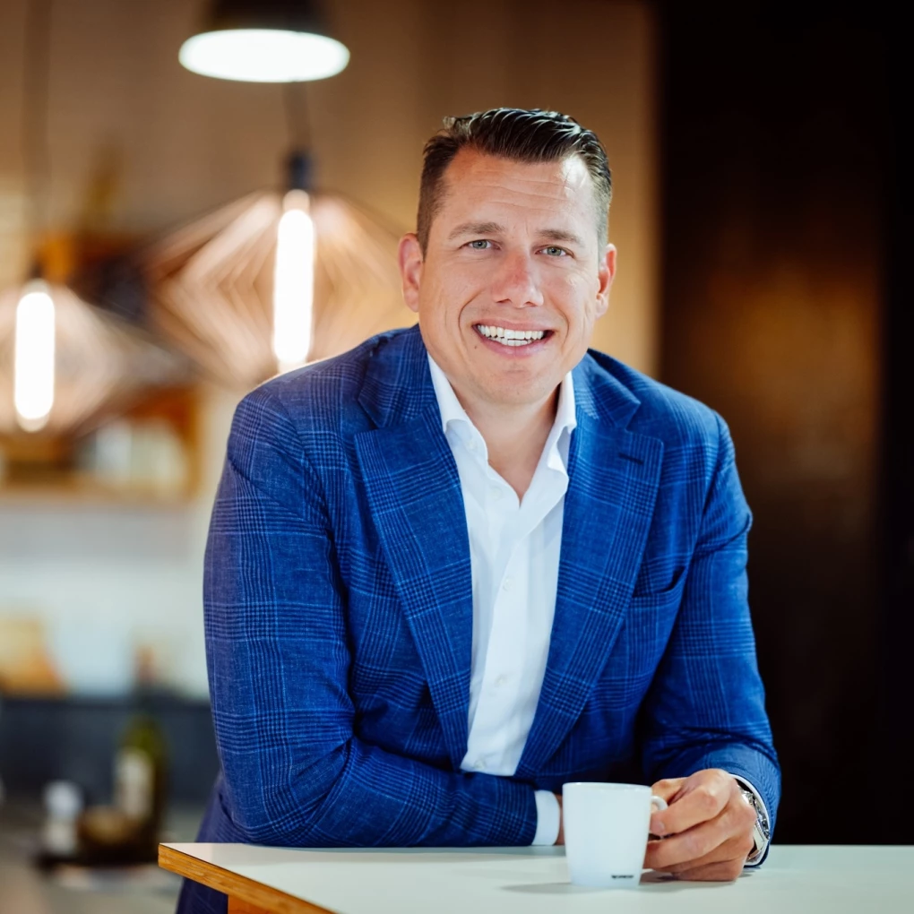 Een man in een blauw pak glimlacht en houdt een kopje koffie vast in een moderne kantooromgeving.