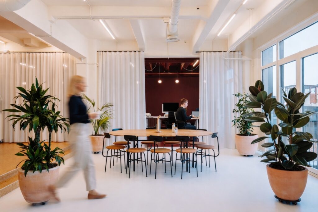 Lichte coworkingruimte met grote tafel en stoelen, omringd door planten, met een persoon aan het werk en een andere die langsloopt.