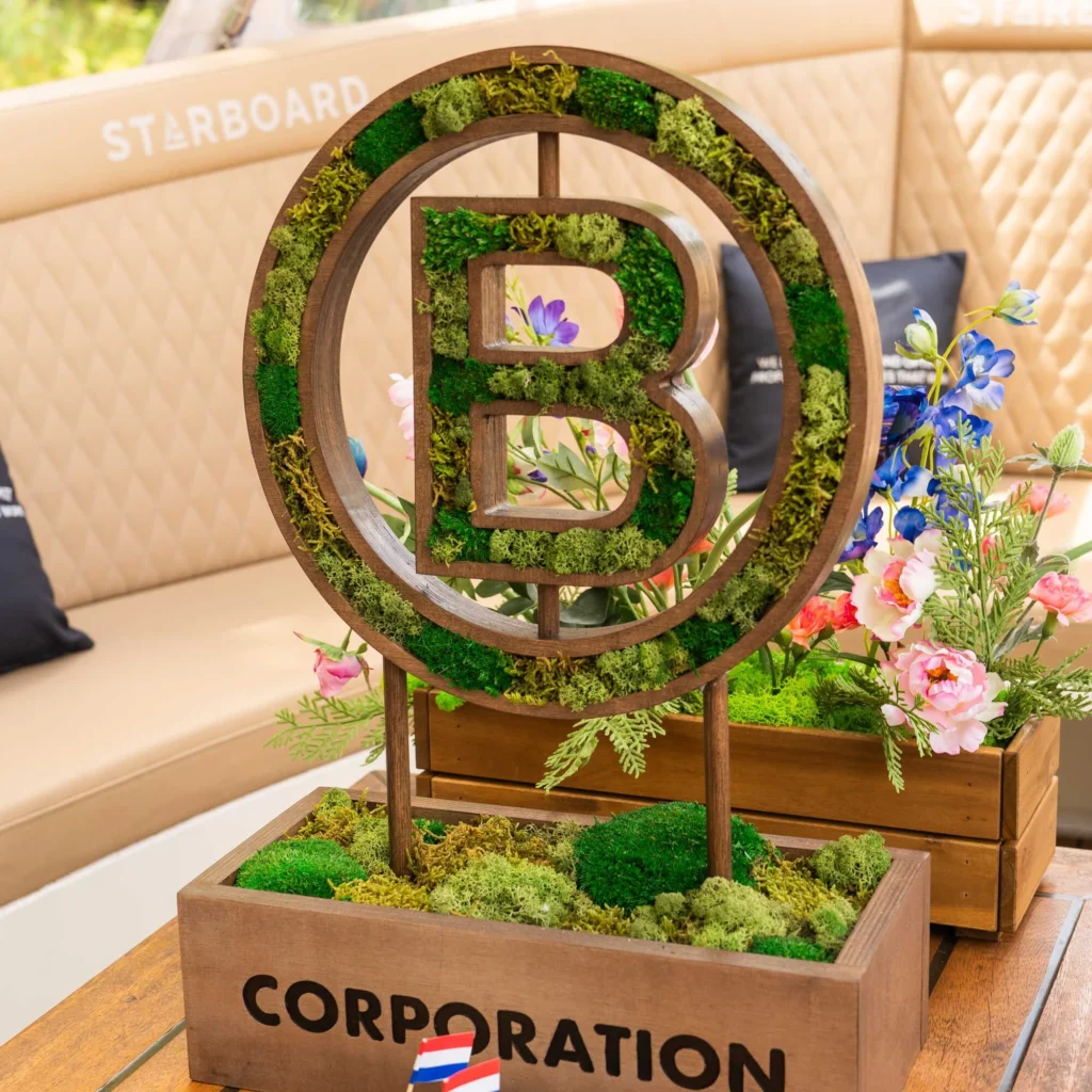 Een houten decoratief object met een 'B' erin, omringd door mos en bloemen, op een tafel met de tekst "Corporation".