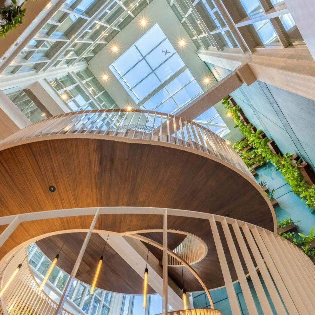 Moderne atriumruimte met houten spiraaltrap, veel glas en groen, waarbij daglicht via het glazen dak naar binnen valt.