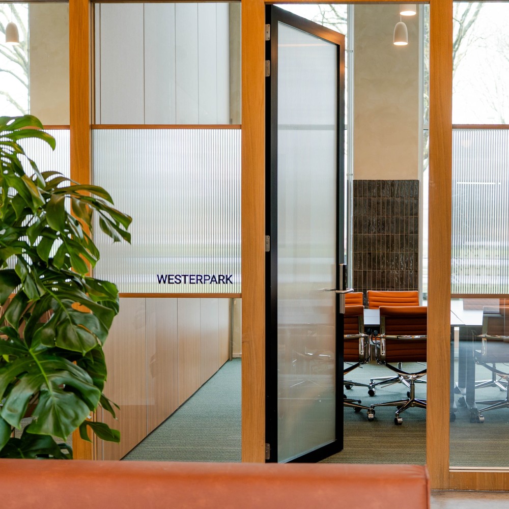 Kantoorinterieur met een glazen deur naar een vergaderruimte, omgeven door planten, met de naam "Westerpark" op de deur.
