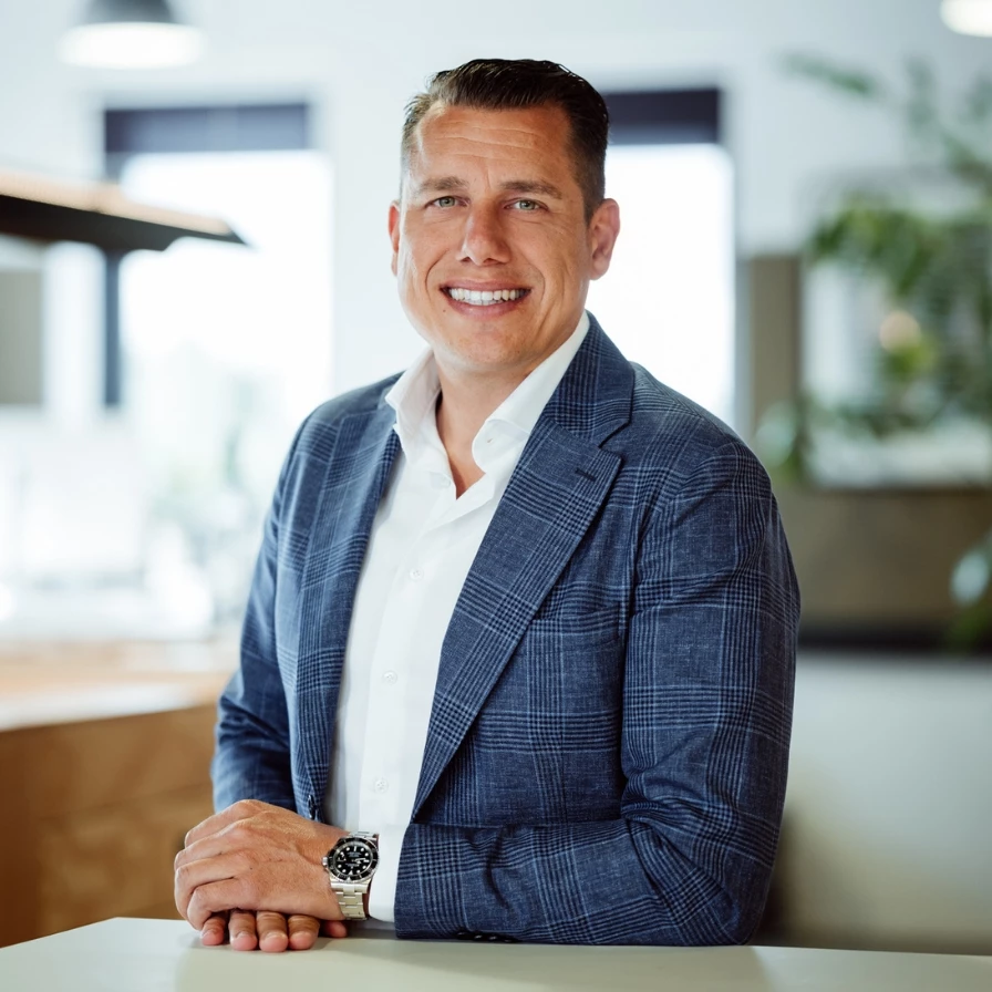 Een man in een zakelijke outfit glimlacht terwijl hij in een moderne kantooromgeving staat.