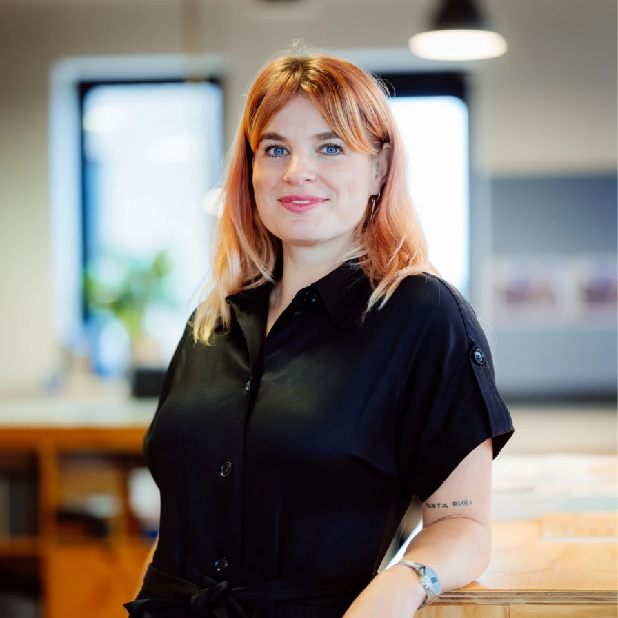 Vrouw met rood haar en een zwart overhemd poseert in een moderne kantooromgeving.