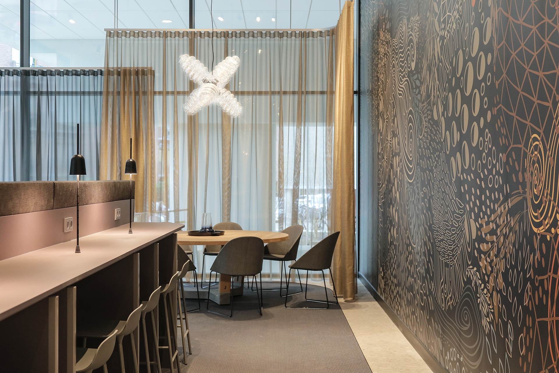 Een moderne vergaderruimte met een hoge bartafel en krukken aan de linkerzijde. Achterin staat een ronde tafel met stoelen bij grote ramen met transparante gordijnen. Aan de rechterkant is een wand met een decoratief patroon zichtbaar en boven de tafel ha