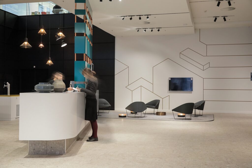 Een moderne lobby met een ronde receptiebalie waar een persoon onscherp aan staat. Rechts bevindt zich een zithoek met lage stoelen en kleine tafeltjes tegen een witte muur met minimalistische lijntekeningen. Het plafond is voorzien van spots en designver