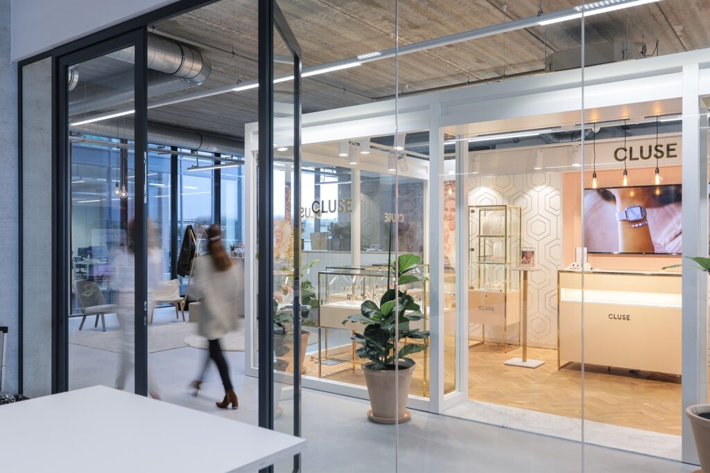 Een moderne kantoorruimte met glazen wanden en een aparte ruimte met de tekst “Exclusive Space” op het glas. Binnen staan een bureau, planten en displays in een lichte inrichting met houten vloer. Links loopt een persoon onscherp door de gang in een open