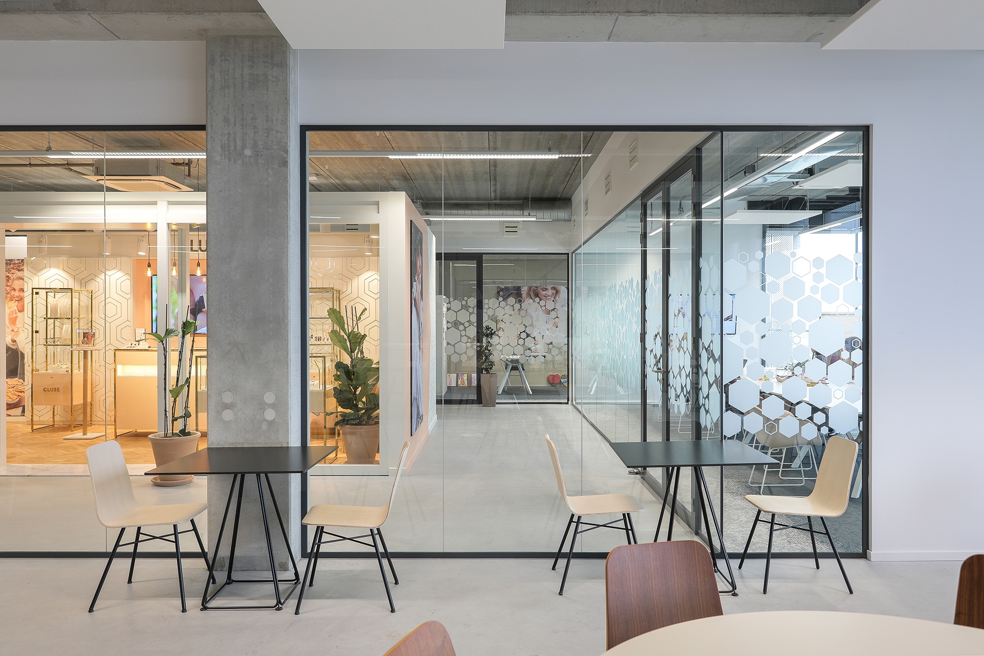 Een moderne kantoorruimte met glazen vergaderruimtes en zwarte frames. Op het glas zijn subtiele patronen zichtbaar. Voor de glazen wanden staan kleine ronde tafels met lichte stoelen. De ruimte heeft een industriële uitstraling met betonnen kolommen en e