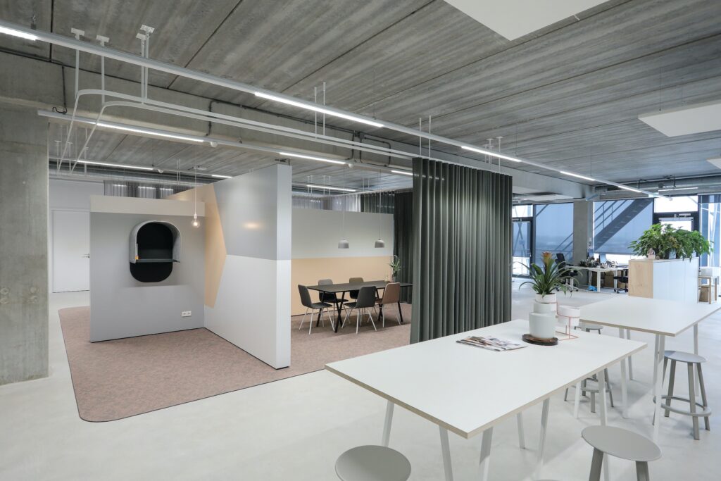 Een modern kantoorinterieur met een industriële uitstraling, betonnen plafond en zichtbare leidingen. In de ruimte staan witte tafels en stoelen, een vergaderruimte met houten tafel en stoelen achter een halfhoge wand, en een groene gordijnwand die de rui