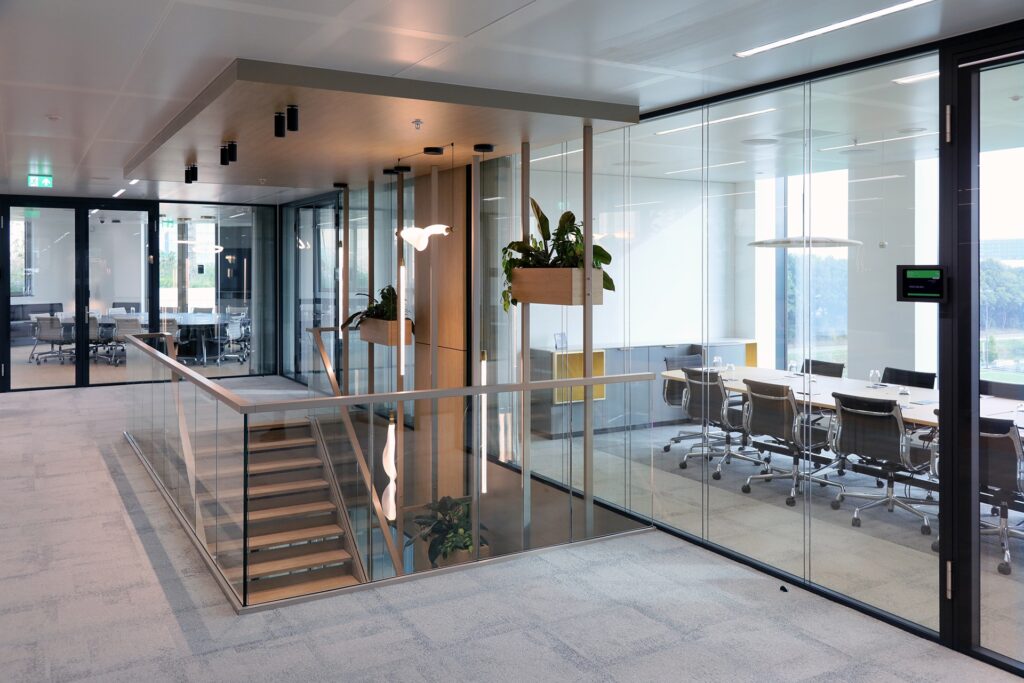 Een moderne kantoorruimte met een open trap en glazen balustrade. Rechts bevindt zich een glazen vergaderruimte met een tafel en stoelen. Planten in houten bakken hangen langs de glazen wanden en grote ramen zorgen voor veel natuurlijk licht.