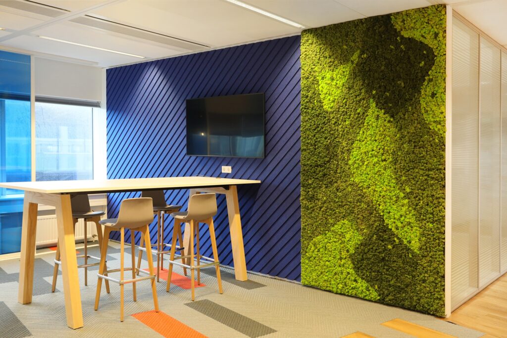 Een moderne kantoorruimte met een hoge tafel en houten barkrukken tegen een blauwe wand met diagonale latten. Aan de muur hangt een flatscreen. Rechts bevindt zich een groene plantenwand met een opvallend patroon. De vloer heeft kleurrijke accenten in ora