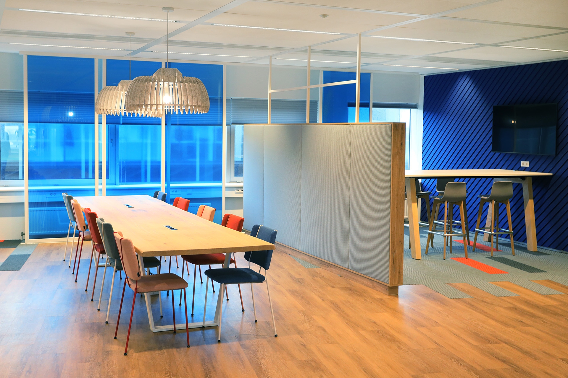 Een moderne kantoorruimte met een lange houten vergadertafel en gekleurde stoelen op een lichte houten vloer. Boven de tafel hangt een grote designlamp. Rechts bevindt zich een hoge tafel met barkrukken tegen een blauwe wand met een flatscreen. Grote rame