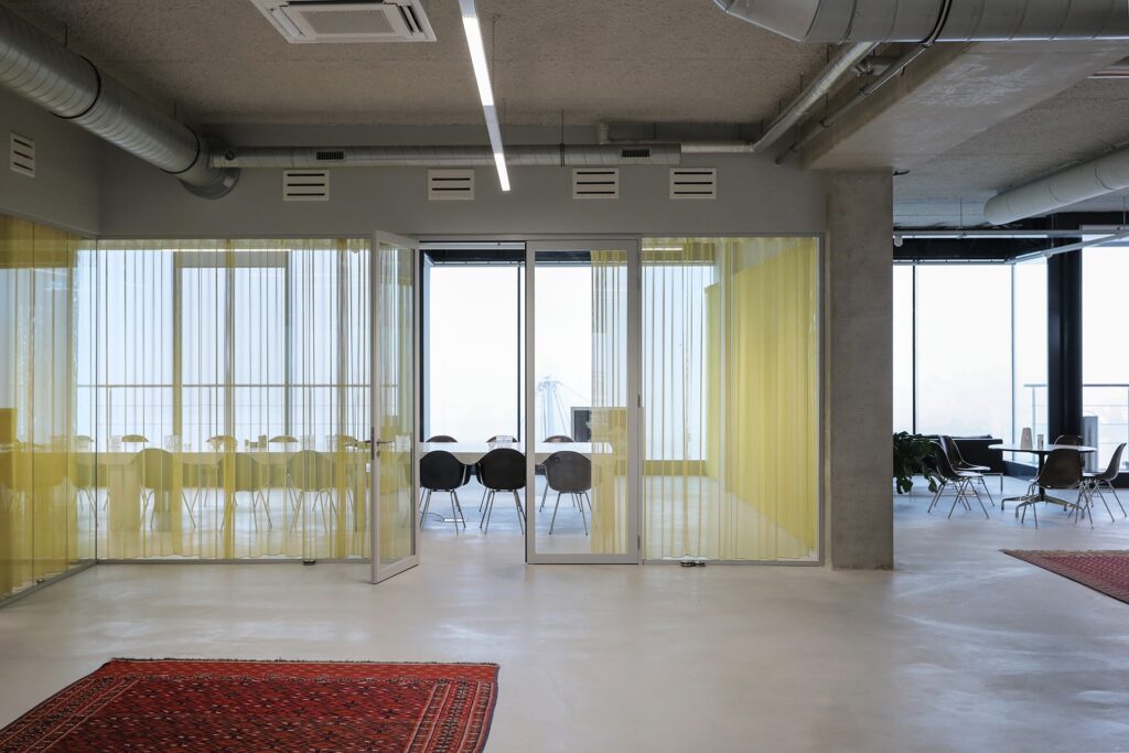 Een moderne kantoorruimte met een glazen vergaderruimte achter gele transparante gordijnen. Binnen staan een tafel en meerdere stoelen. De ruimte heeft een industriële uitstraling met betonnen kolommen, zichtbare ventilatiebuizen en een strakke vloer.