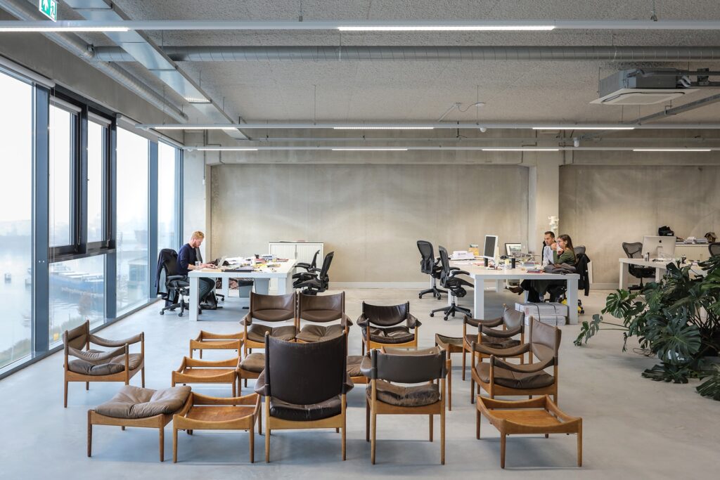 Moderne open kantoorruimte met meerdere bureaus en medewerkers aan het werk, met op de voorgrond rijen houten stoelen en grote ramen met uitzicht op de stad.