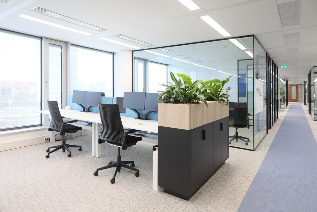 Een moderne kantoorruimte met meerdere bureaus en zwarte bureaustoelen bij grote ramen. In het midden staat een kast met een houten bovenkant en groene planten. Rechts bevindt zich een glazen vergaderruimte met zwarte frames. De ruimte is licht en strak i