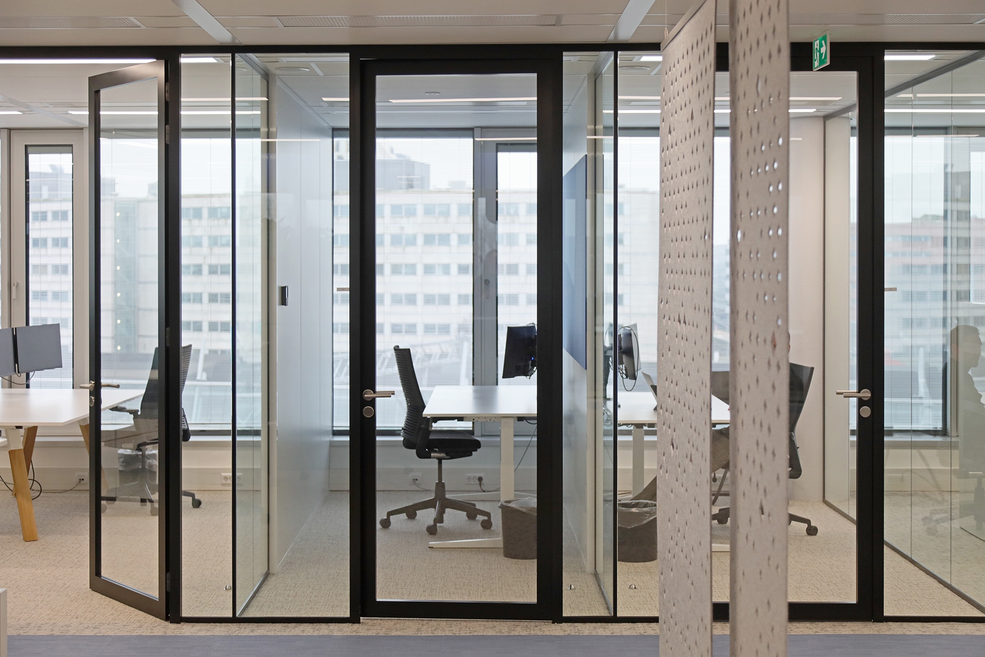 Een moderne kantoorruimte met glazen vergader- en werkruimtes met zwarte frames. Binnen staan bureaus met bureaustoelen bij grote ramen met uitzicht op andere gebouwen. Op de voorgrond is een houten wand met een geperforeerd patroon zichtbaar.