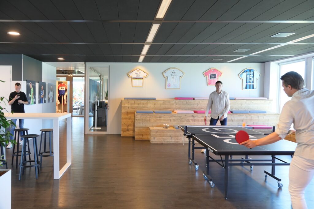 Twee collega’s spelen tafeltennis in een moderne kantoorruimte met houten balie, donkere vloer en ingelijste sportshirts aan de muur.