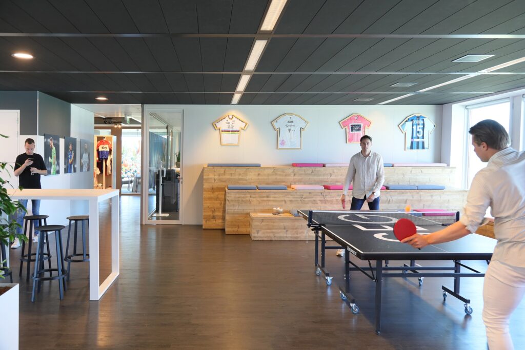 Een moderne kantoorruimte met een tafeltennistafel op de voorgrond waar twee personen aan spelen. Aan de muur hangen ingelijste sportshirts boven een houten tribune met zitplaatsen. De ruimte heeft een donkere vloer en een strak plafond met verlichting.