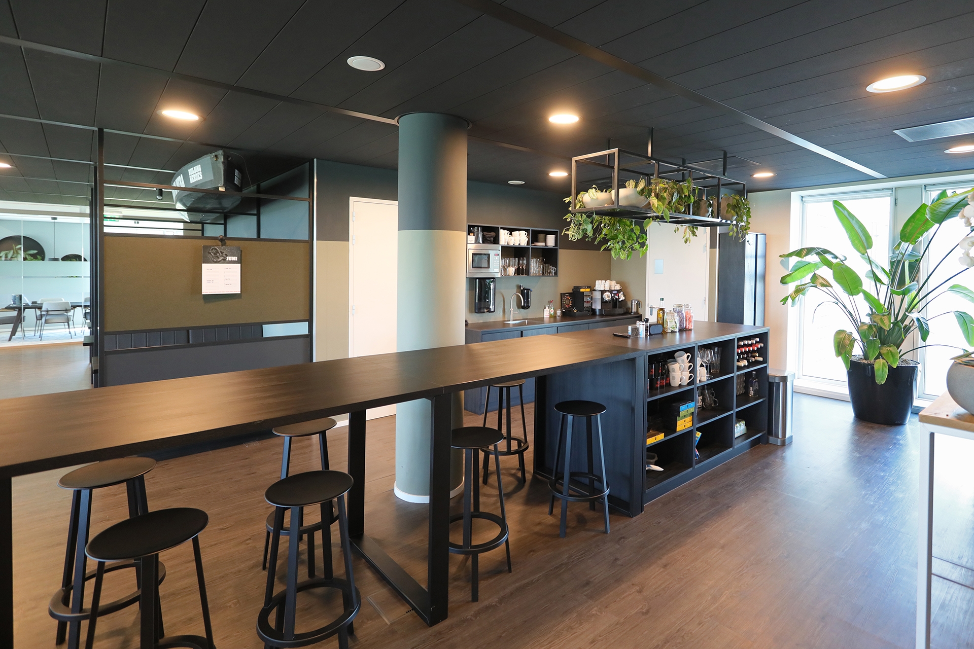 Een moderne kantoorpantry met een lange hoge tafel en zwarte barkrukken. Achter de bar staan koffiemachines, glazen en flessen op open planken. Planten hangen boven de bar en grote ramen zorgen voor veel natuurlijk licht in de ruimte.