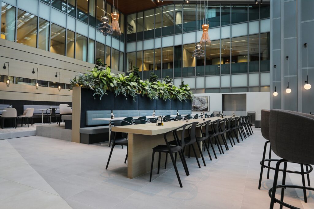 Een moderne kantoorruimte met een lange eettafel en zwarte stoelen in een open atrium. Achter de tafel staat een hoge plantenbak met groene beplanting en een ingebouwde zitbank. Boven de ruimte hangen designlampen en rondom zijn glazen gevels zichtbaar va