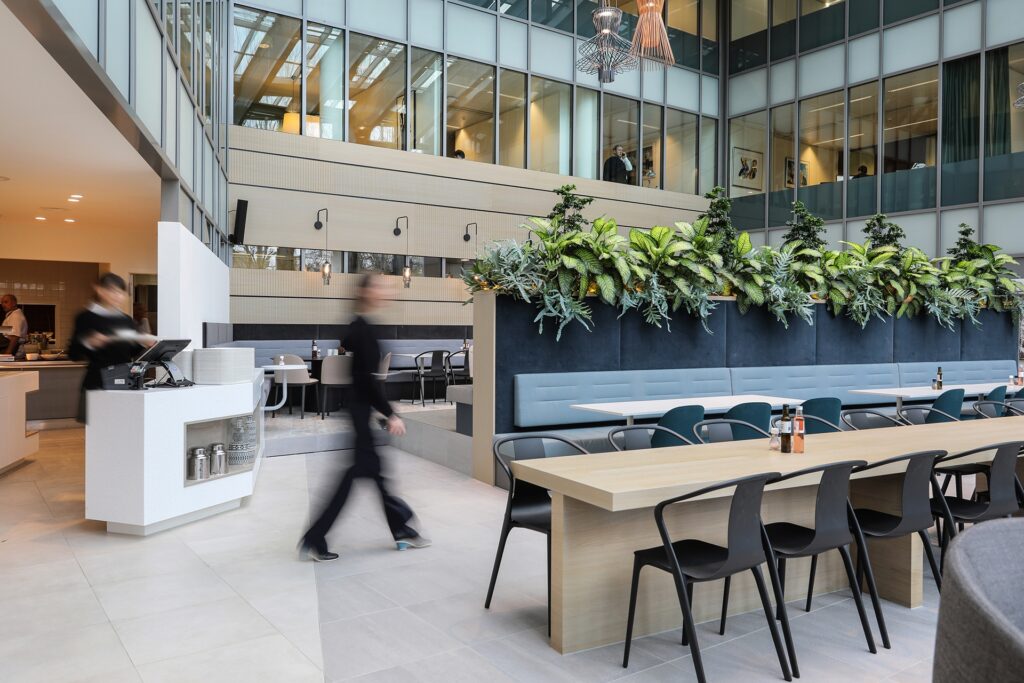 Een moderne kantoorruimte met een lange houten tafel en zwarte stoelen in een open atrium. Achter de tafel staat een hoge plantenbak met groene beplanting en een ingebouwde zitbank. Links bevindt zich een koffiecorner en een persoon loopt onscherp door he