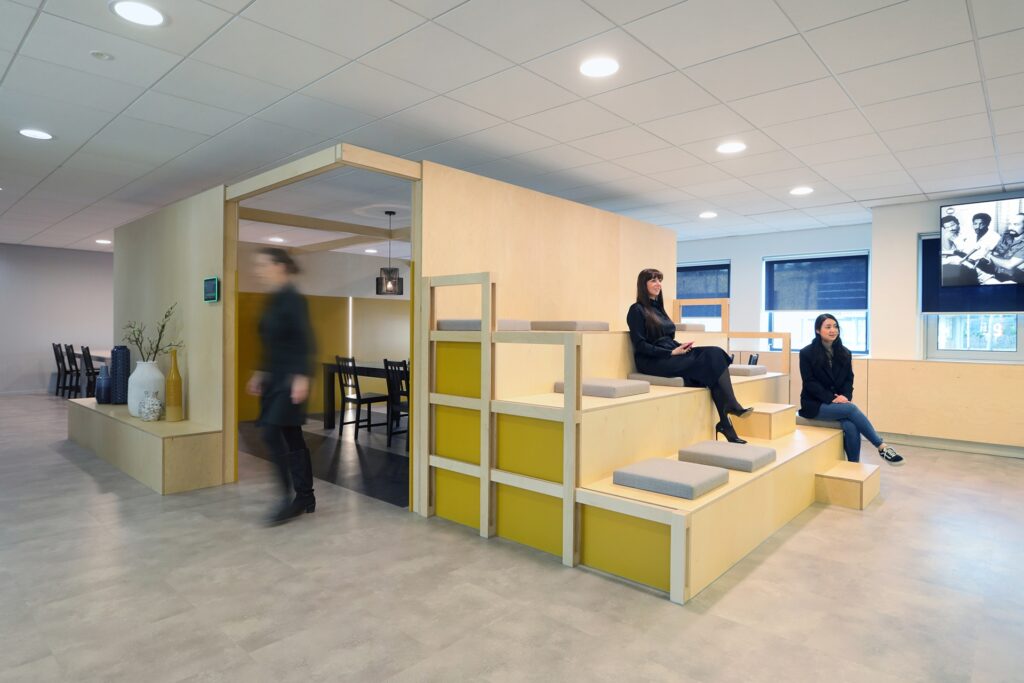 Een moderne kantoorruimte met een houten tribuneconstructie met geïntegreerde zitplaatsen en opbergvakken in geel. Twee personen zitten op de treden en praten, terwijl links een persoon onscherp voorbijloopt. Op de achtergrond is een vergaderruimte met ta