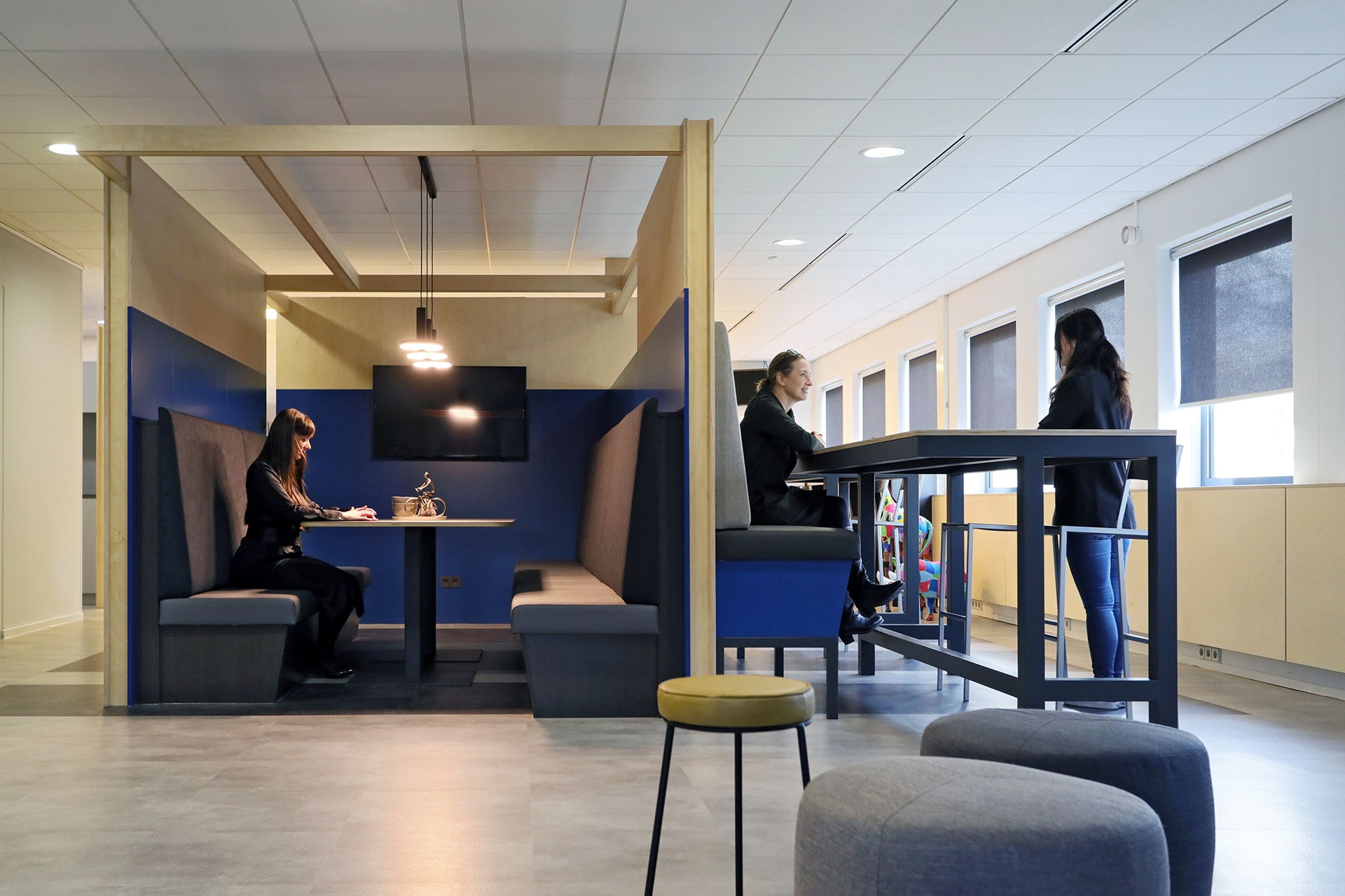 Een moderne kantoorruimte met twee verschillende overlegplekken. Links zit een persoon in een afgesloten zithoek met blauwe wanden aan een tafel. Rechts staan twee personen aan een hoge tafel met barkrukken bij een raam. Op de voorgrond staan ronde poefje